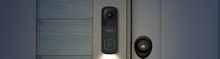 A black Lorex 4K Wi-Fi video doorbell with a green-lit button, mounted beside a front door. The doorbell emits a soft white light, providing visibility in low-light conditions, and features a sleek, modern design.