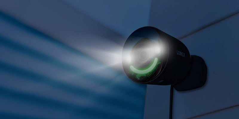 A black 4K outdoor security camera with a spotlight and green smart security lighting, mounted on a wall. The camera emits a bright white light, designed for enhanced visibility and nighttime security, with the brand logo "Lorex" visible on its side.