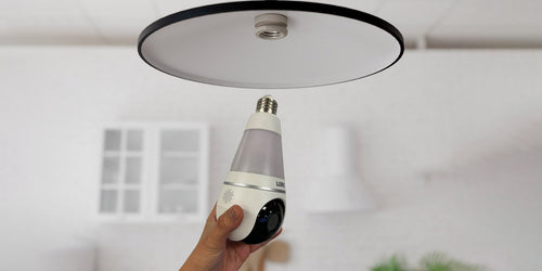 A person prepares to install a Lorex 2K Wi-Fi lightbulb security camera into an indoor ceiling socket, highlighting its simple screw-in setup and built-in camera lens.